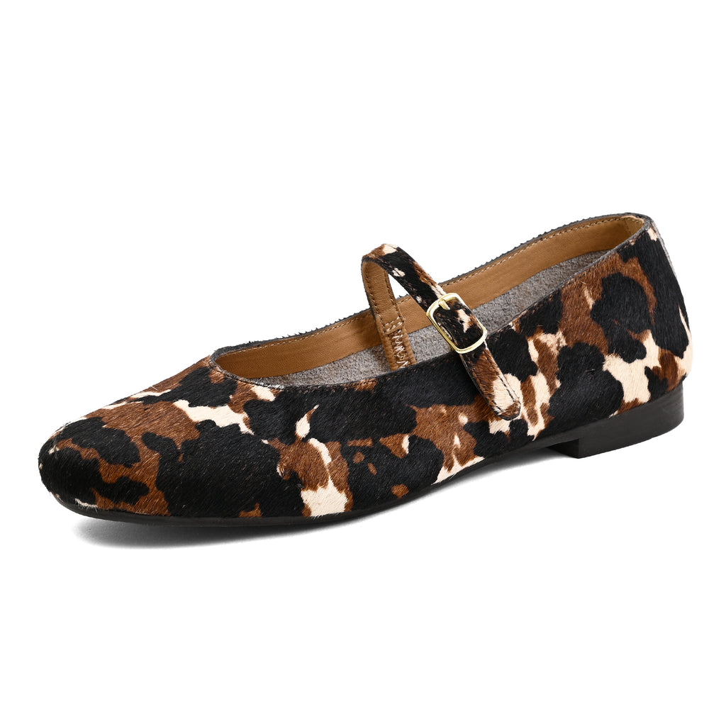 A single women's Mary Jane flat shoe with a brown, black, and beige cow print pattern, featuring a strap with a gold-tone buckle, shown on a plain white background.